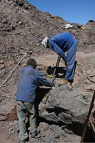 Abb. zeigt zwei Arbeiter im blauer Arbeitskleidung die sich mit Brechstangen abmühen, dem Felsen Kristalle zu entlocken.
